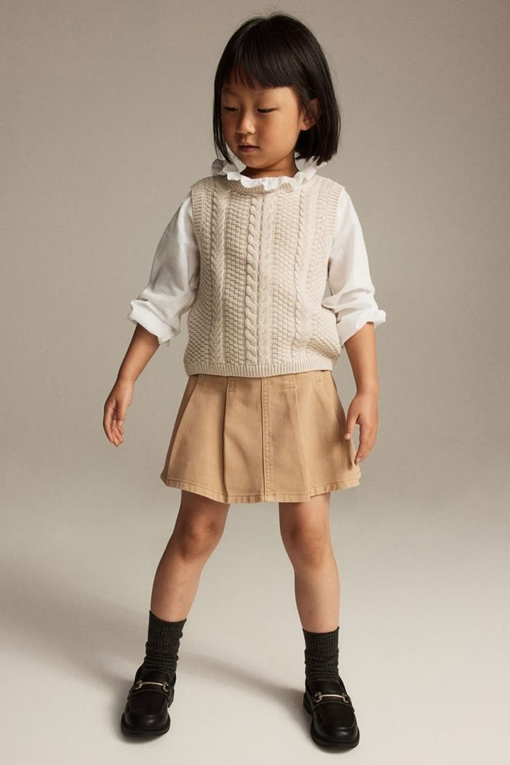 Young child in a white cable knit sweater, khaki pleated skirt, and black shoes photographed in different poses.