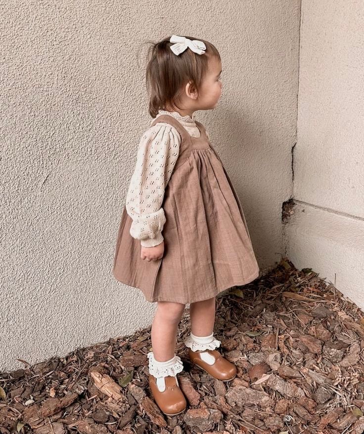 Toddler girl in floral blouse, brown pinafore dress, ruffle socks, and Mary Jane shoes with a white bow hair clip.