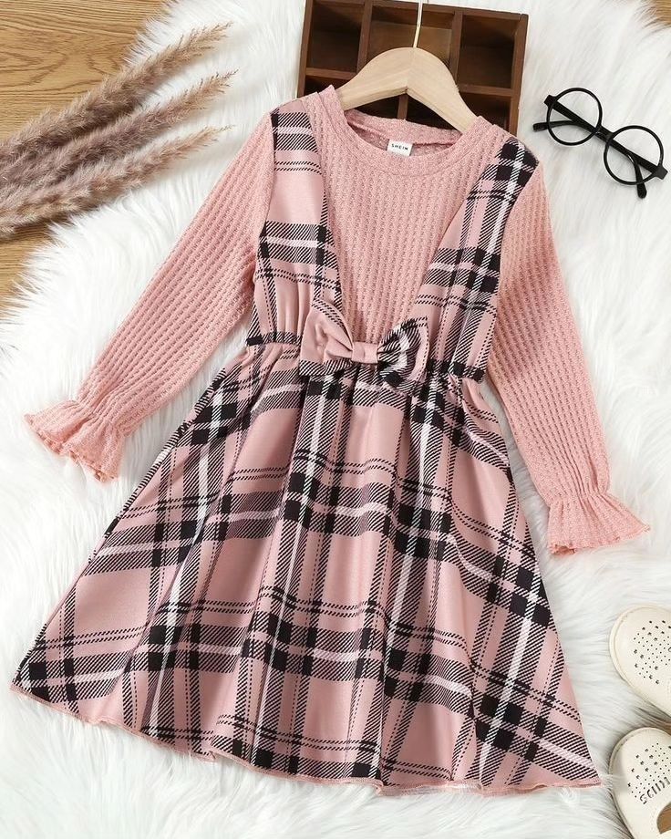 Pink plaid girls dress with long knit sleeves and bow waist detail, displayed on hanger with glasses and shoes.