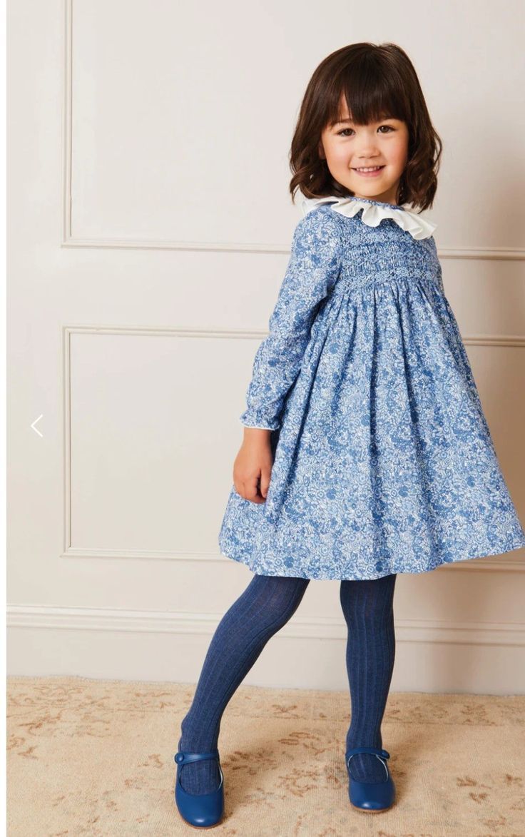 Young girl wearing a blue floral smocked dress with white collar, blue ribbed tights, and blue Mary Jane shoes.