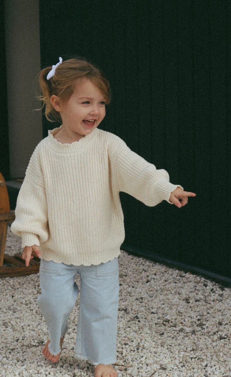 A cheerful toddler girl in a cream knit sweater and light jeans points while smiling outdoors.