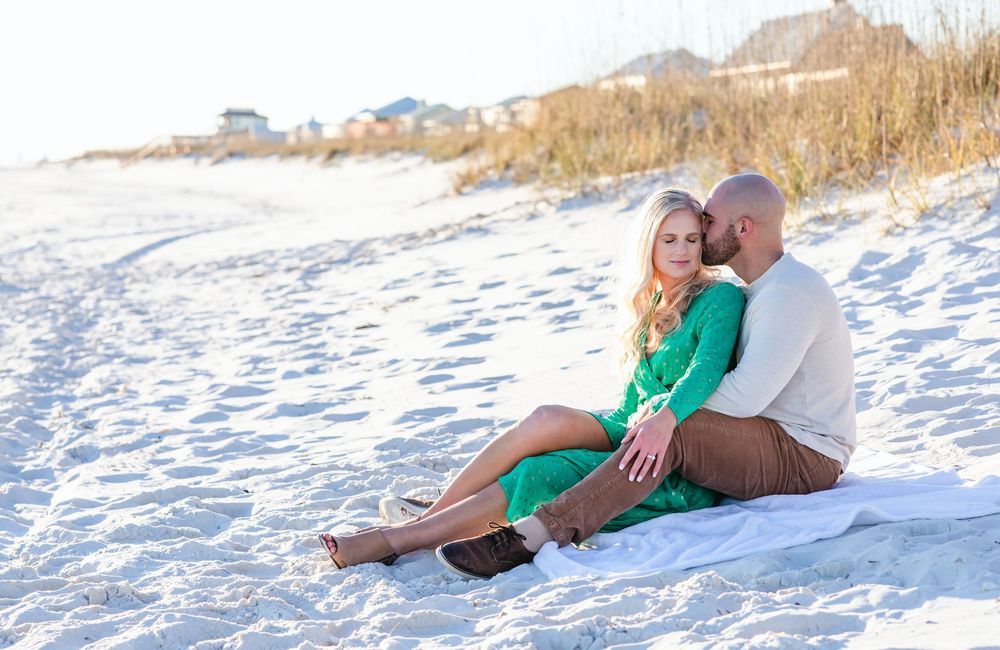 A couple in casual attire sits together on white sandy beach during golden hour.
