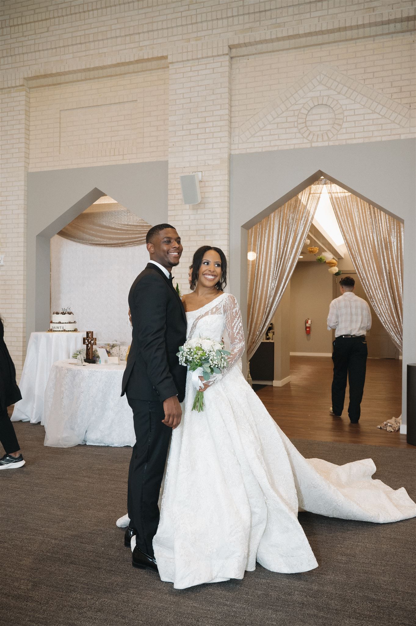 A newlywed couple poses for a photo in an elegant reception venue with draped fabric decor and a cake table nearby.