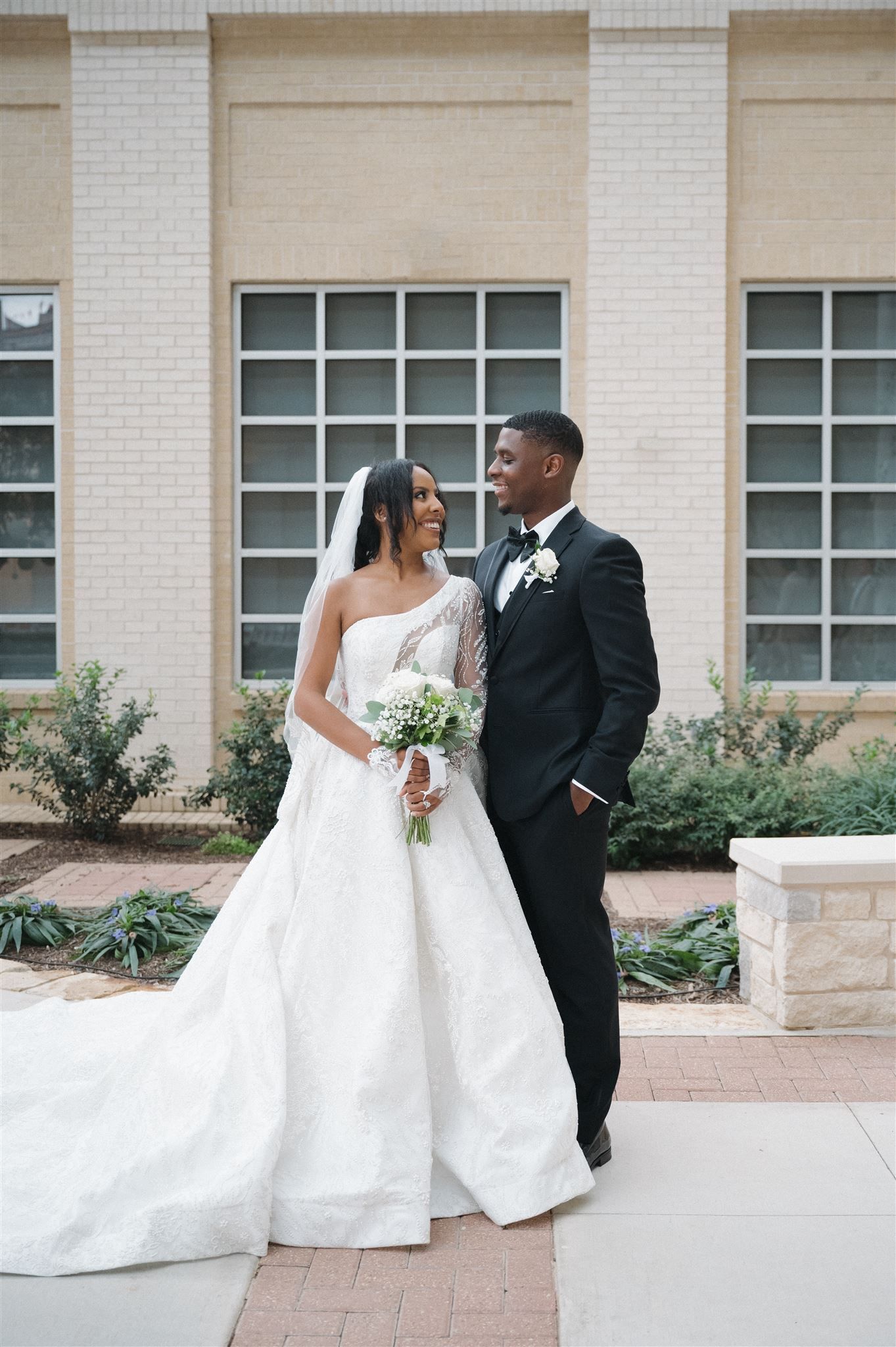 A bride and groom share a loving moment on their wedding day outside a building with white brick walls.