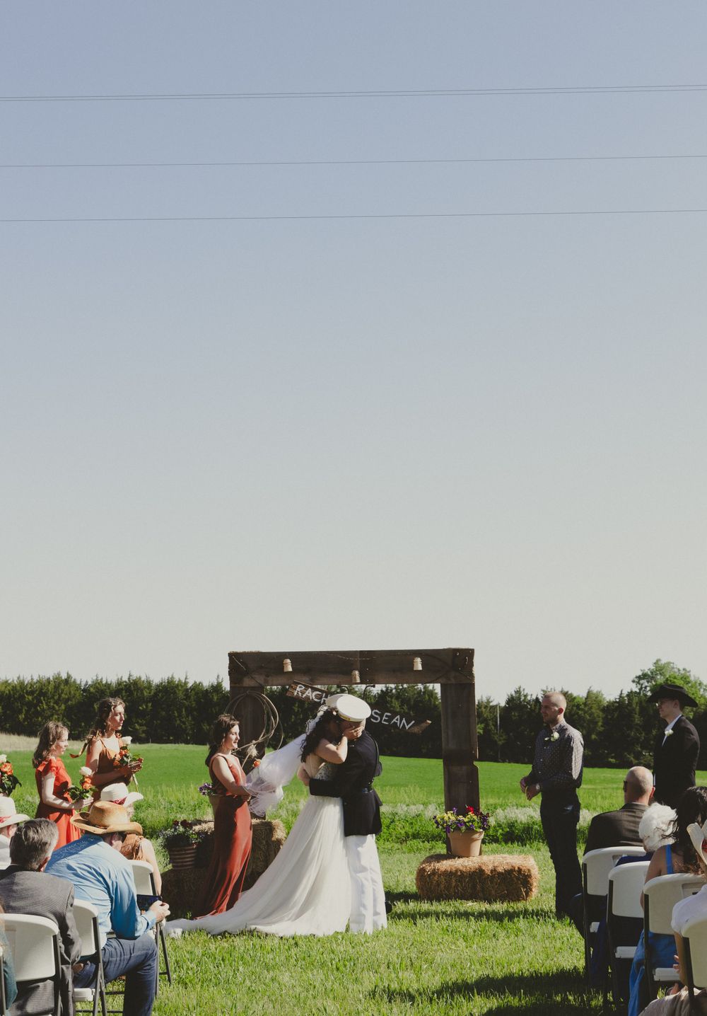 Rustic outdoor wedding ceremony with hay bales and wooden arch in a green field.