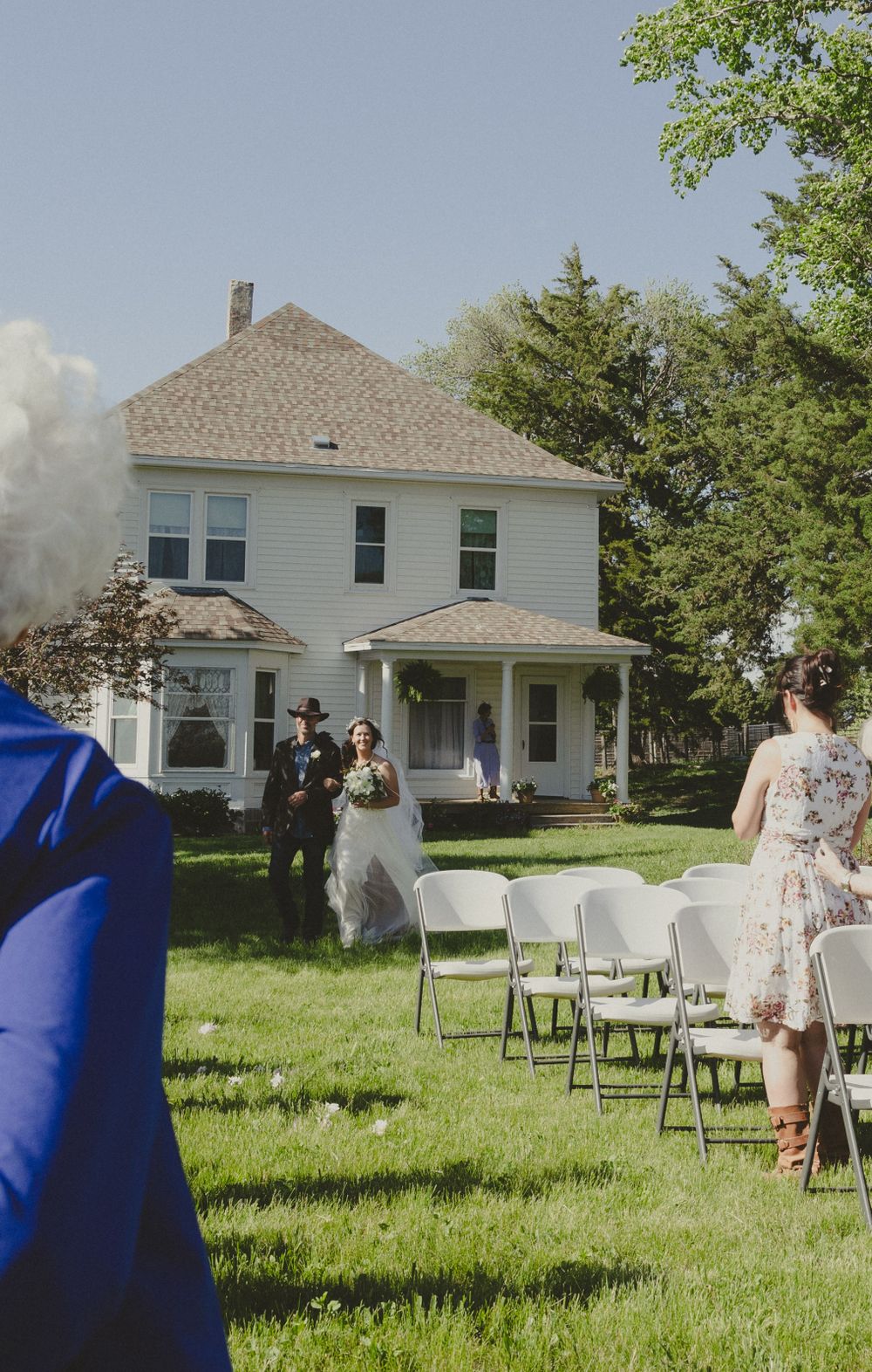 Outdoor wedding setup with white chairs on lawn in front of a classic farmhouse.