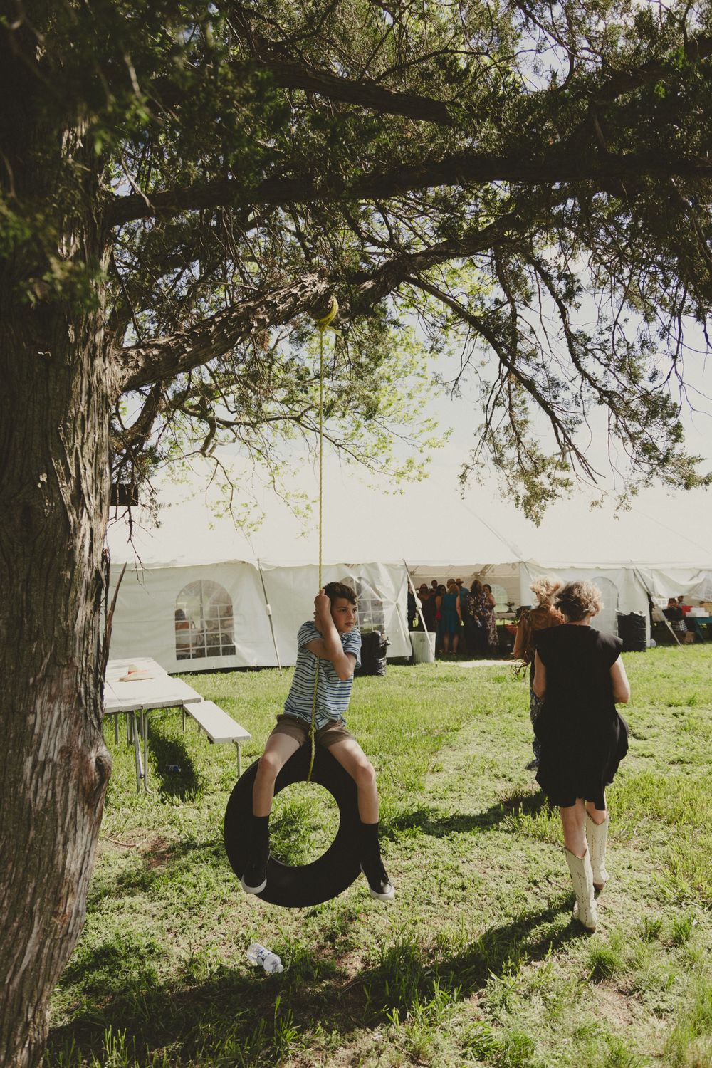Child playing on a tire swing while being photographed under a large shady tree near a white tent.