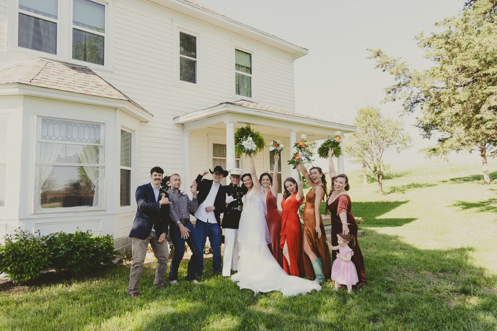 Wedding party poses together outside a white farmhouse with green hanging plants on a sunny day.