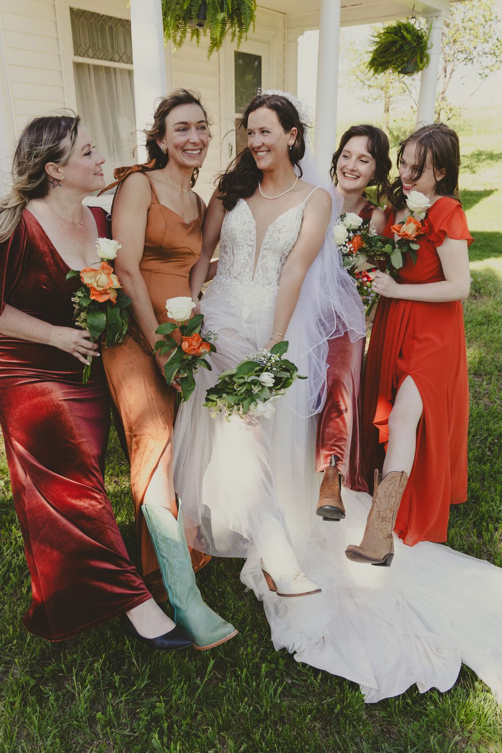 Bridesmaids in colorful dresses laugh together on a white porch with bouquets.