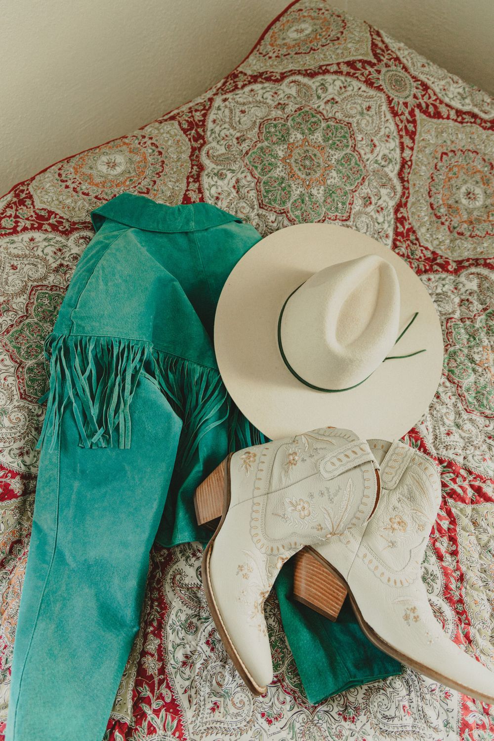 Western-style outfit layout with turquoise pants, white cowboy boots and cream cowboy hat on patterned fabric.