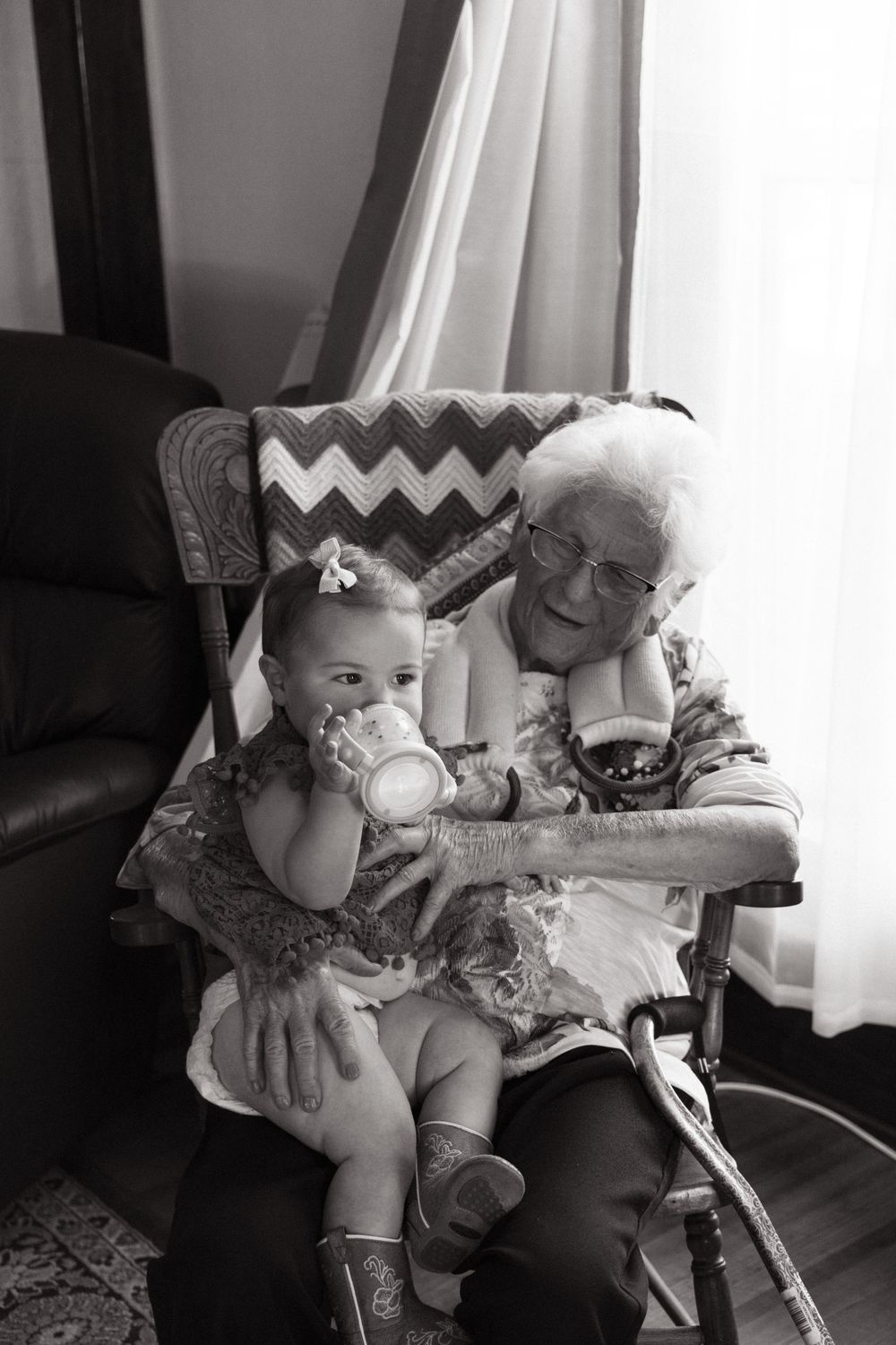 Black and white photo of an elderly person and baby sitting together in a chair reading a book.