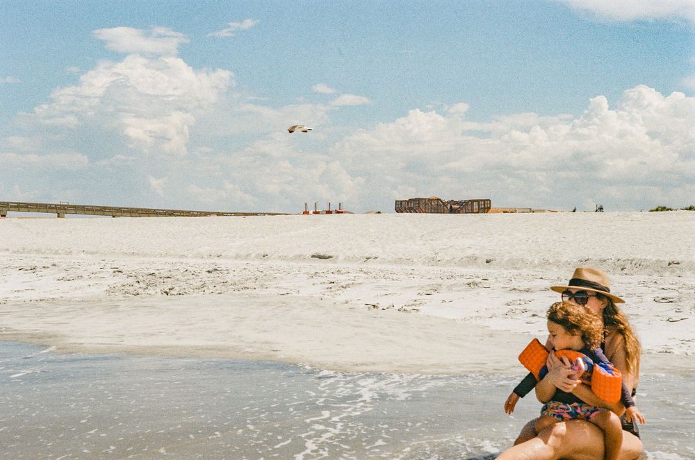 A tranquil beach scene with white sand and distant buildings under a blue summer sky.