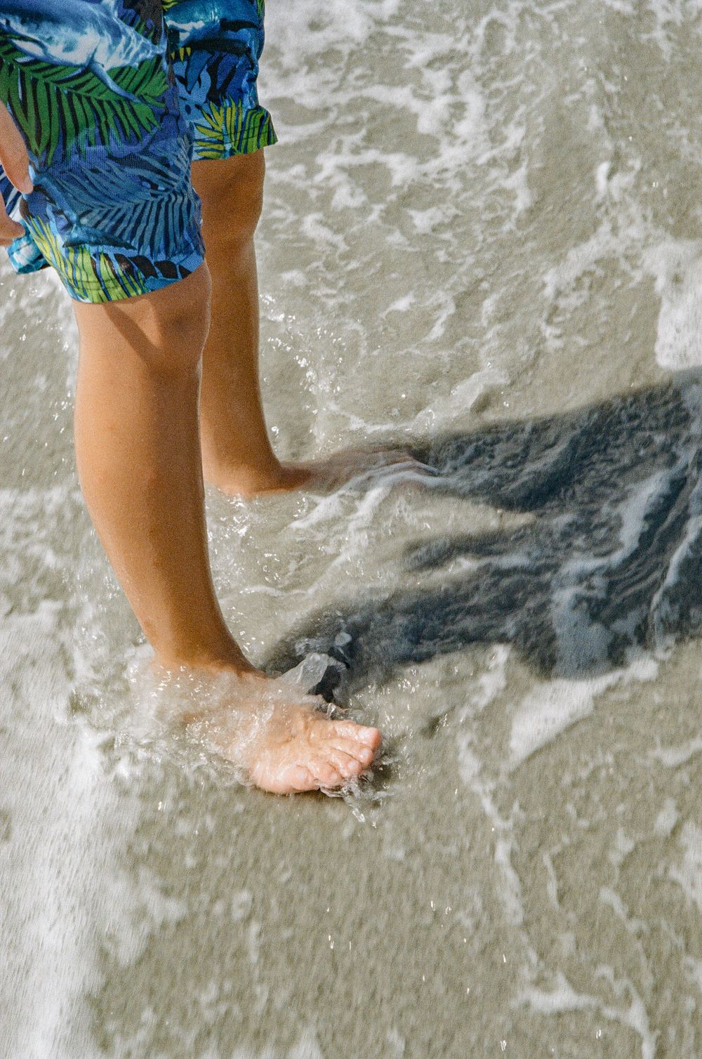 Feet in tropical print swim shorts standing in ocean surf.
