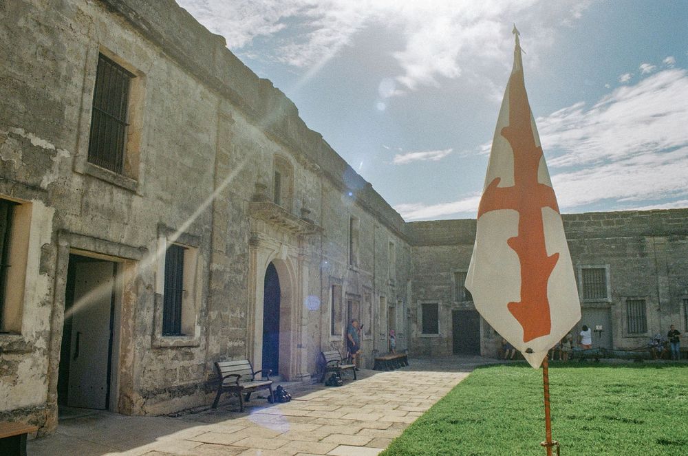 Historic stone fortress courtyard with blue door and orange flag under cloudy sky.