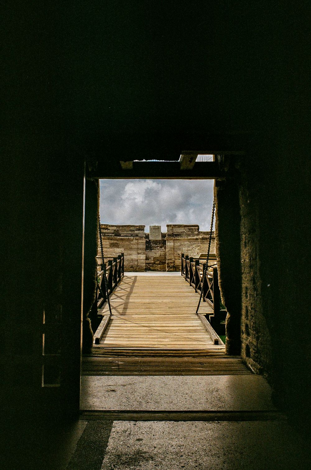 Dark stone passageway opening to a wooden boardwalk under cloudy skies.