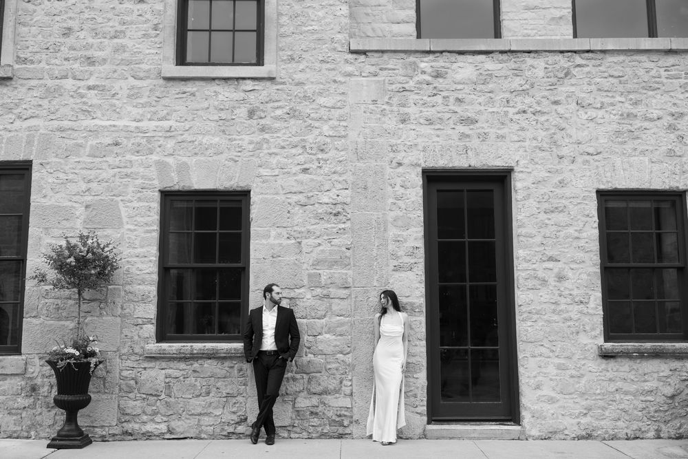 A couple in formal attire poses against a rustic stone wall featuring tall windows and a black doorway.