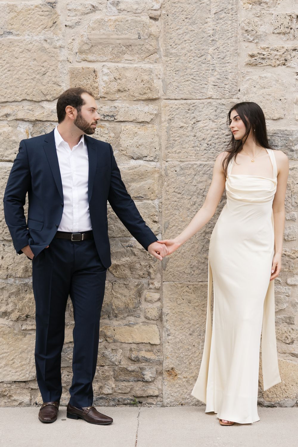 Couple in formal attire hold hands against stone wall, with groom in navy suit and bride in flowing cream gown.