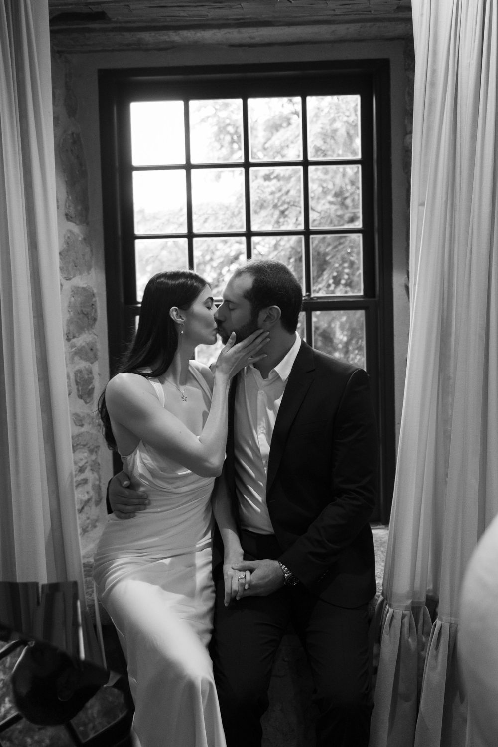 A romantic black and white photo of a couple sharing an intimate moment by a window with curtains and natural light.
