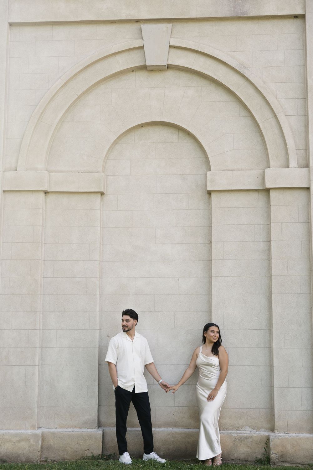 Couple holds hands while standing against a white stone archway wall in elegant minimalist outfits.