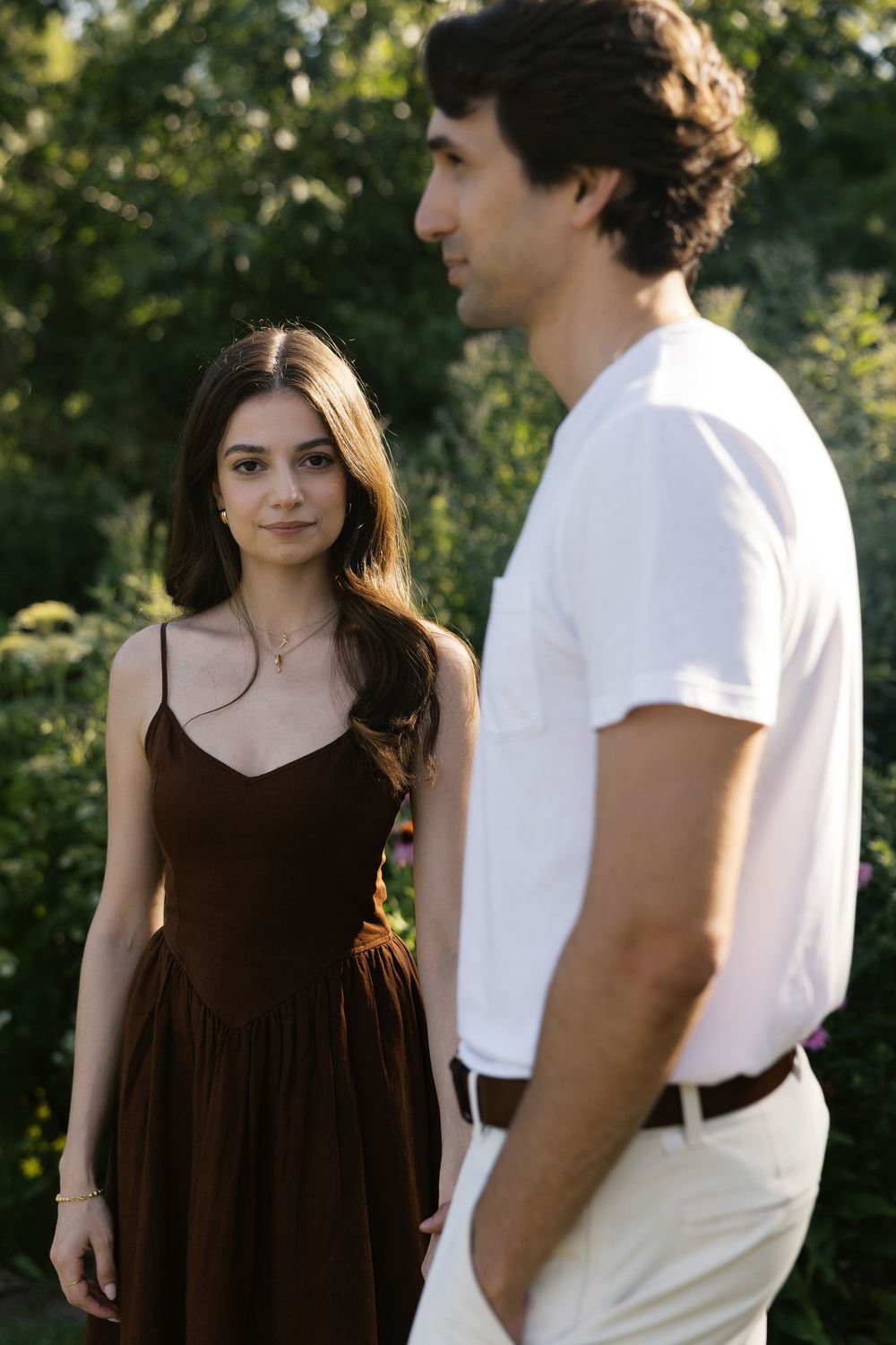 A couple outdoors with one person wearing a brown dress and another in white casual attire amid greenery.