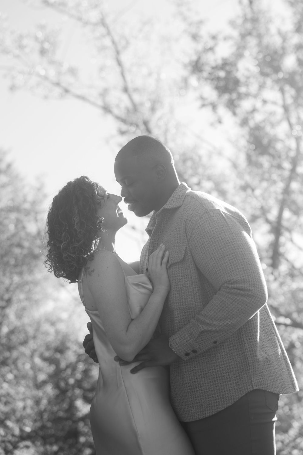 A romantic couple embraces in silhouette against a soft, dreamy background with trees and sunlight.