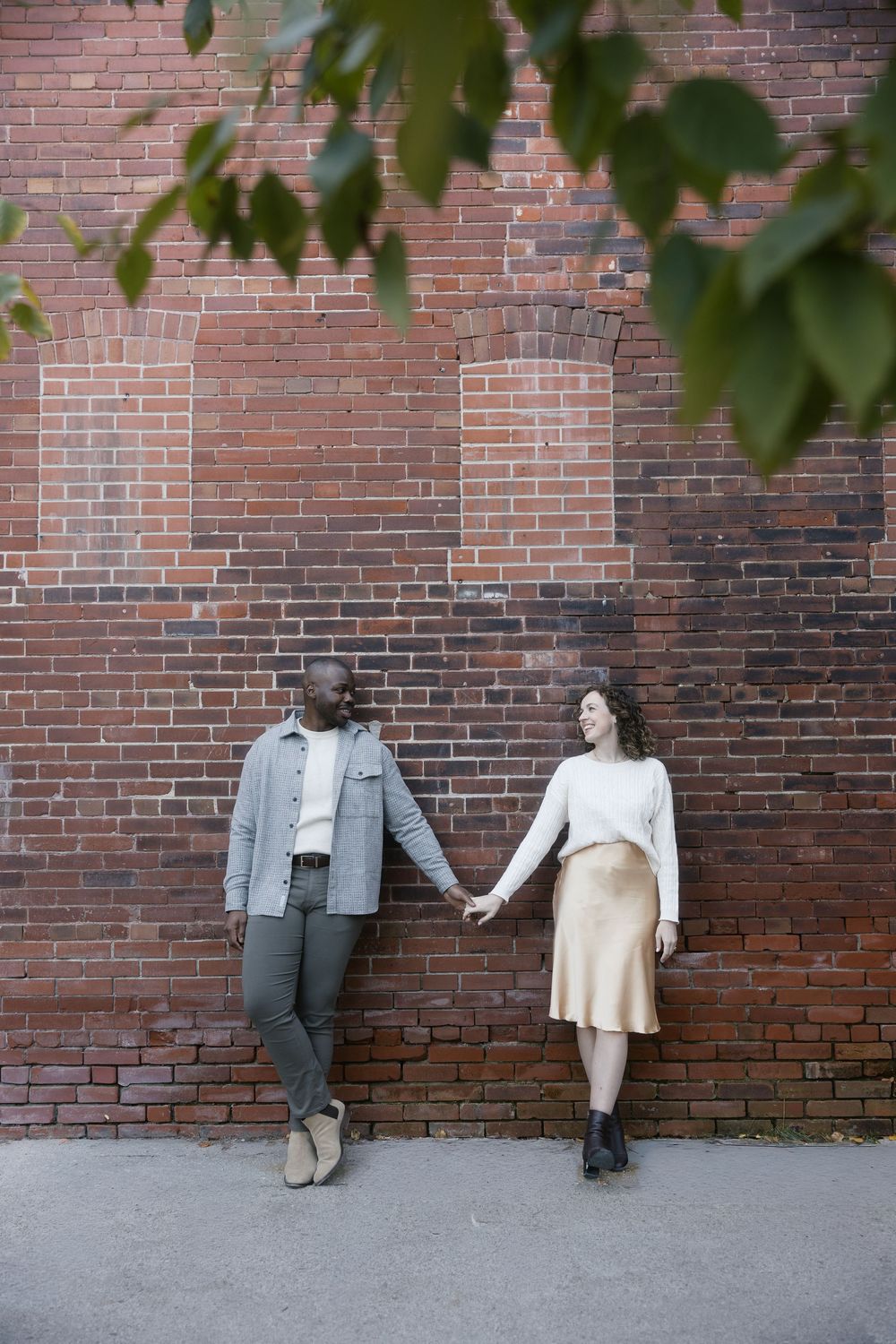 Two mannequins dressed in casual clothing sit against a weathered brick wall under green leaves.
