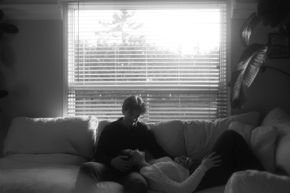 Black and white photo of two people relaxing on a couch near a window with blinds in a cozy living room.