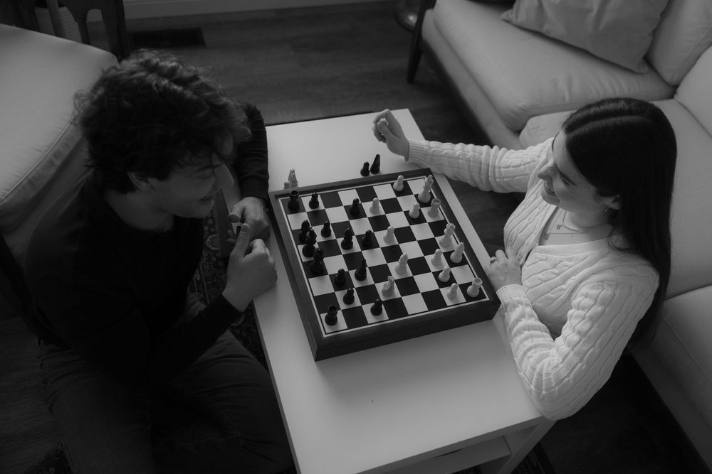 Two people playing chess at a coffee table in a living room, captured in black and white from above.