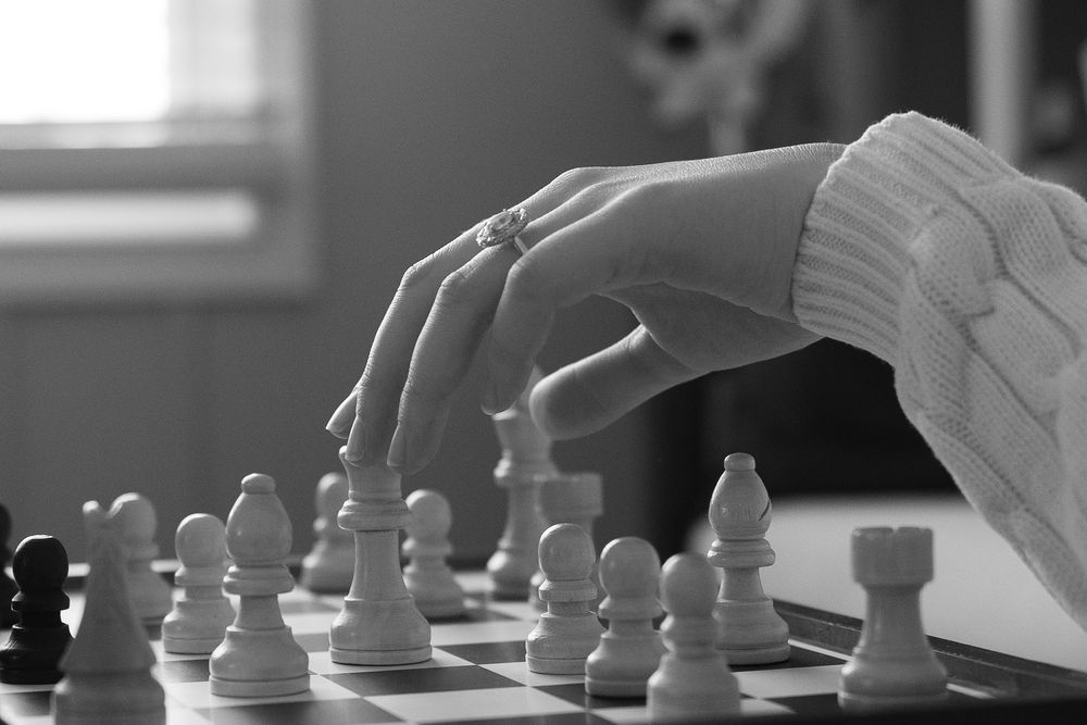 Black and white photo of a hand moving a chess piece on a board during a strategic game.