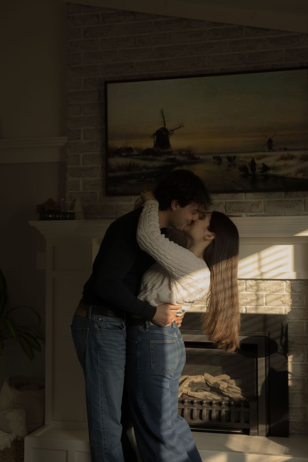 A couple shares a romantic kiss near a fireplace in a warmly lit living room with a windmill painting above.