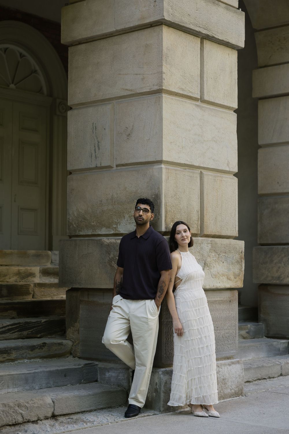 A man in black shirt and a woman in white dress pose elegantly beside a large stone column of a classical building.