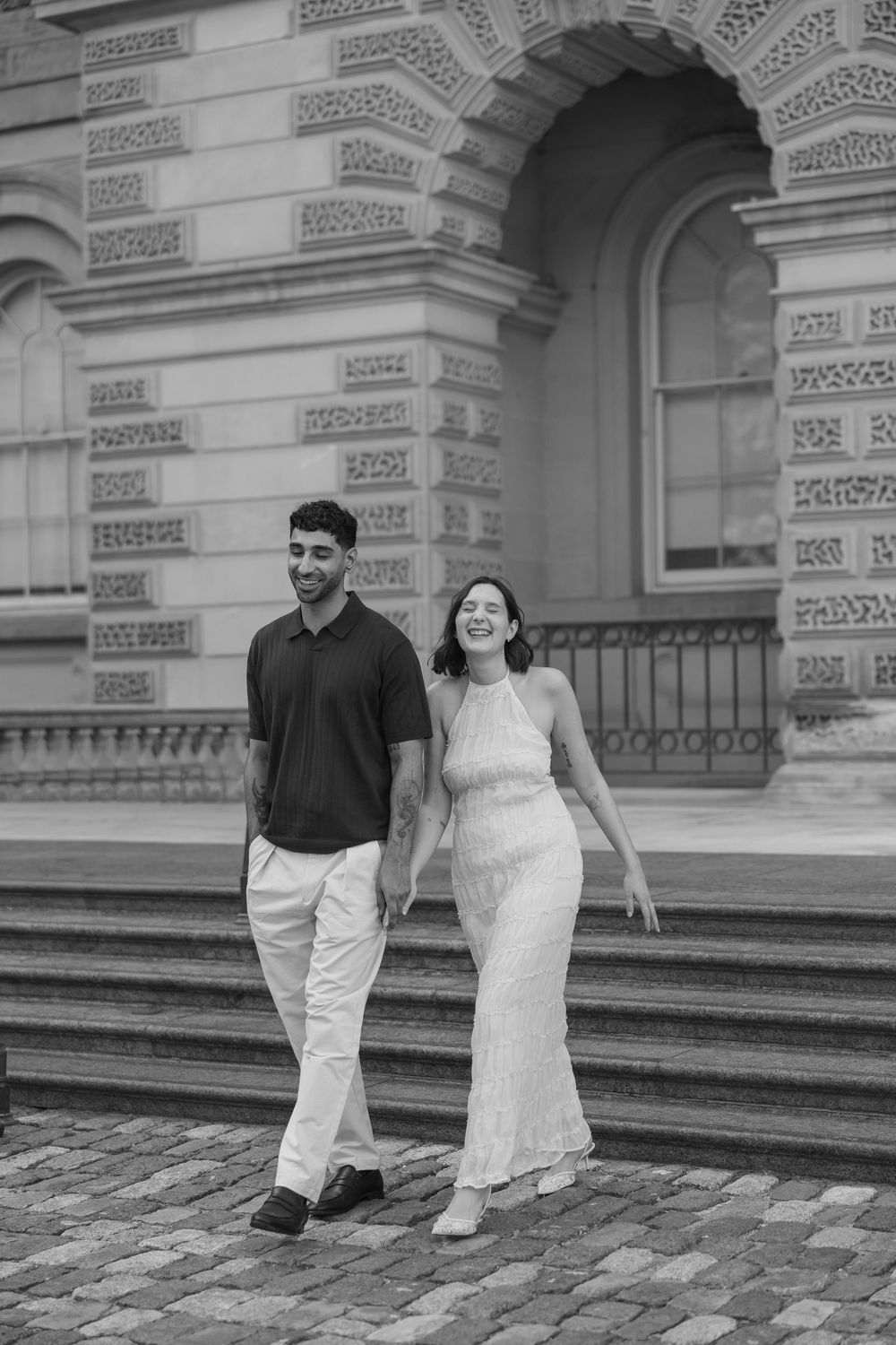 Black and white photo of a smiling couple holding hands walking down steps in front of a grand historic building.