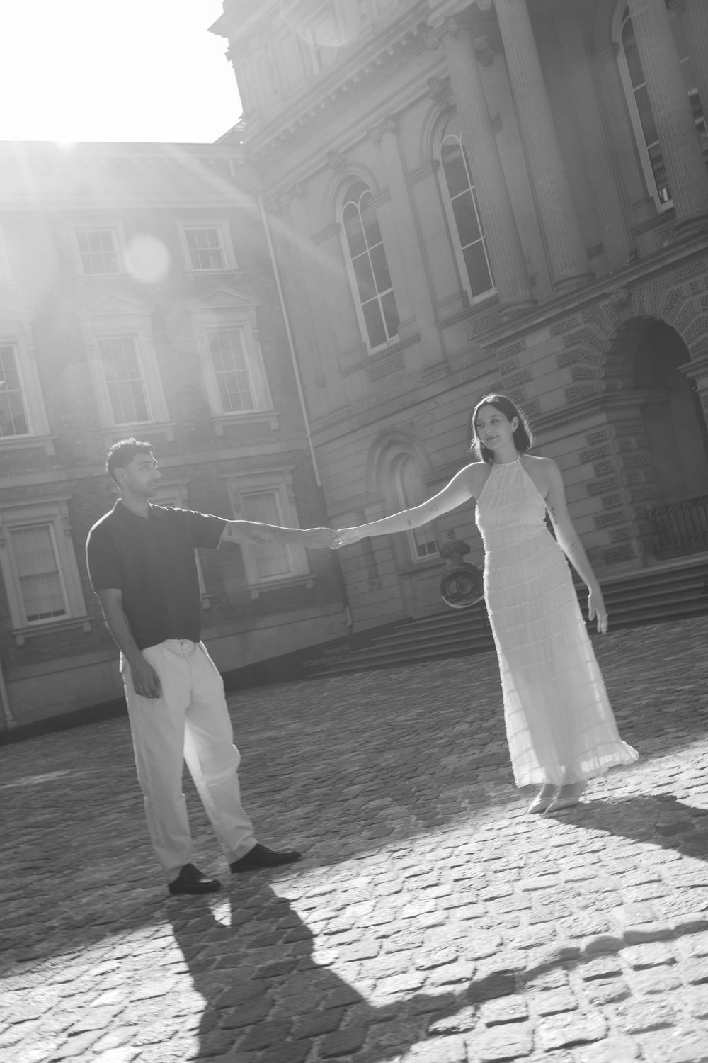 Black and white photo of a couple holding hands on cobblestone plaza in front of a grand historic building in bright sunlight.