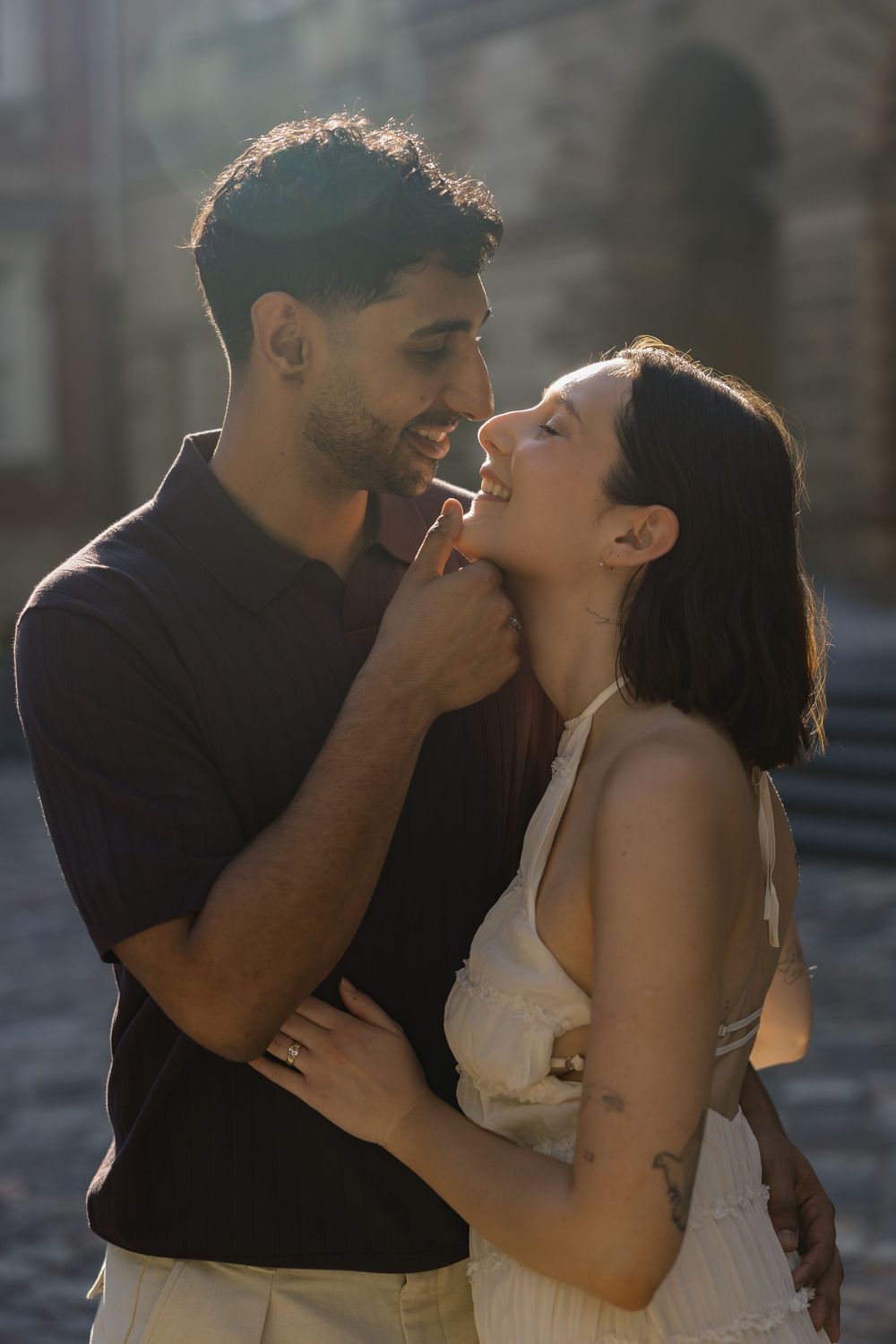 A romantic couple shares an intimate moment outdoors, bathed in warm golden sunlight on a cobblestone street.