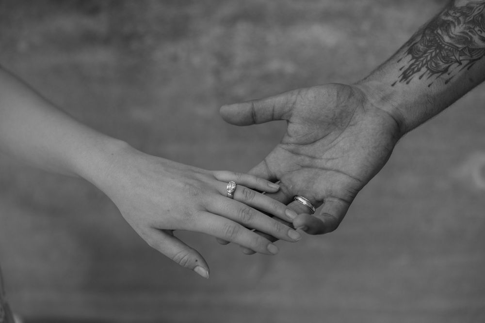 Black and white photo of two hands almost touching, one with a ring and tattoo, symbolizing love and connection.
