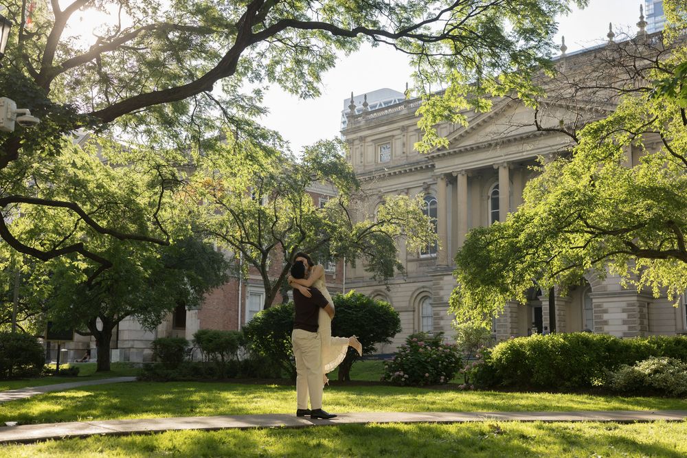 Person standing in front of a grand neoclassical building surrounded by lush green trees on a sunny day.