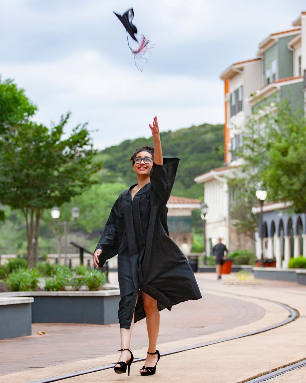 Graduate in black gown tossing cap in celebratory pose on campus walkway.