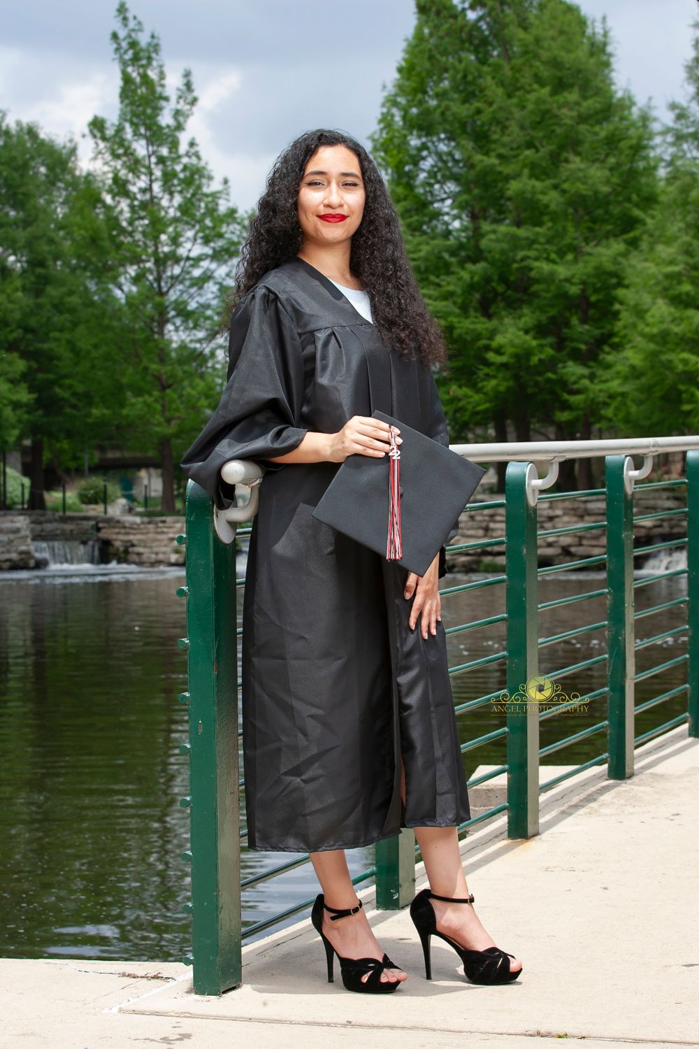 Graduate in black gown posing by waterfront railing with tassel and diploma.