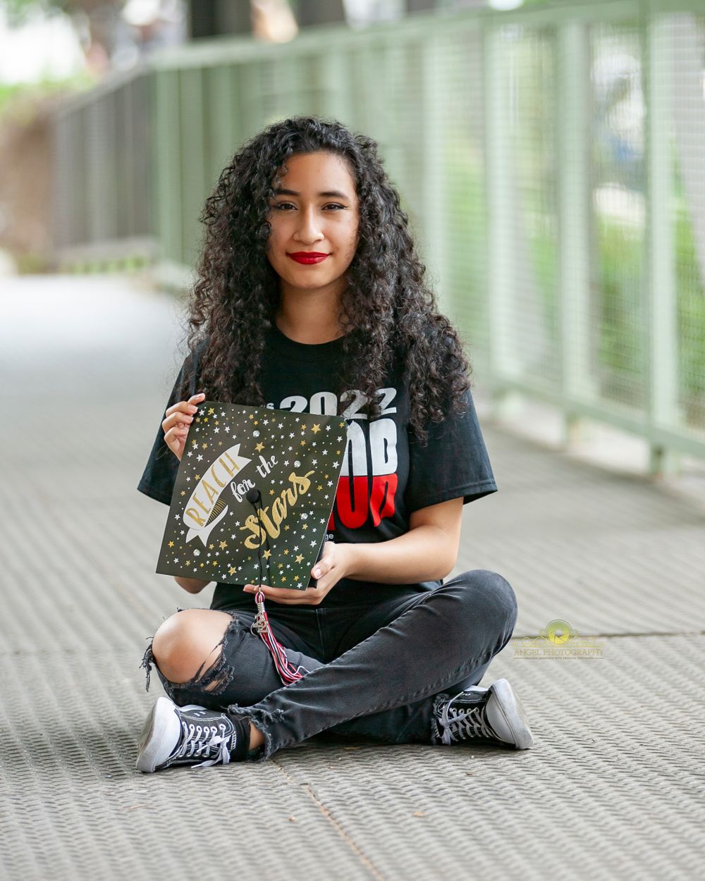Graduate posing with decorated cap while sitting cross-legged on metal walkway.