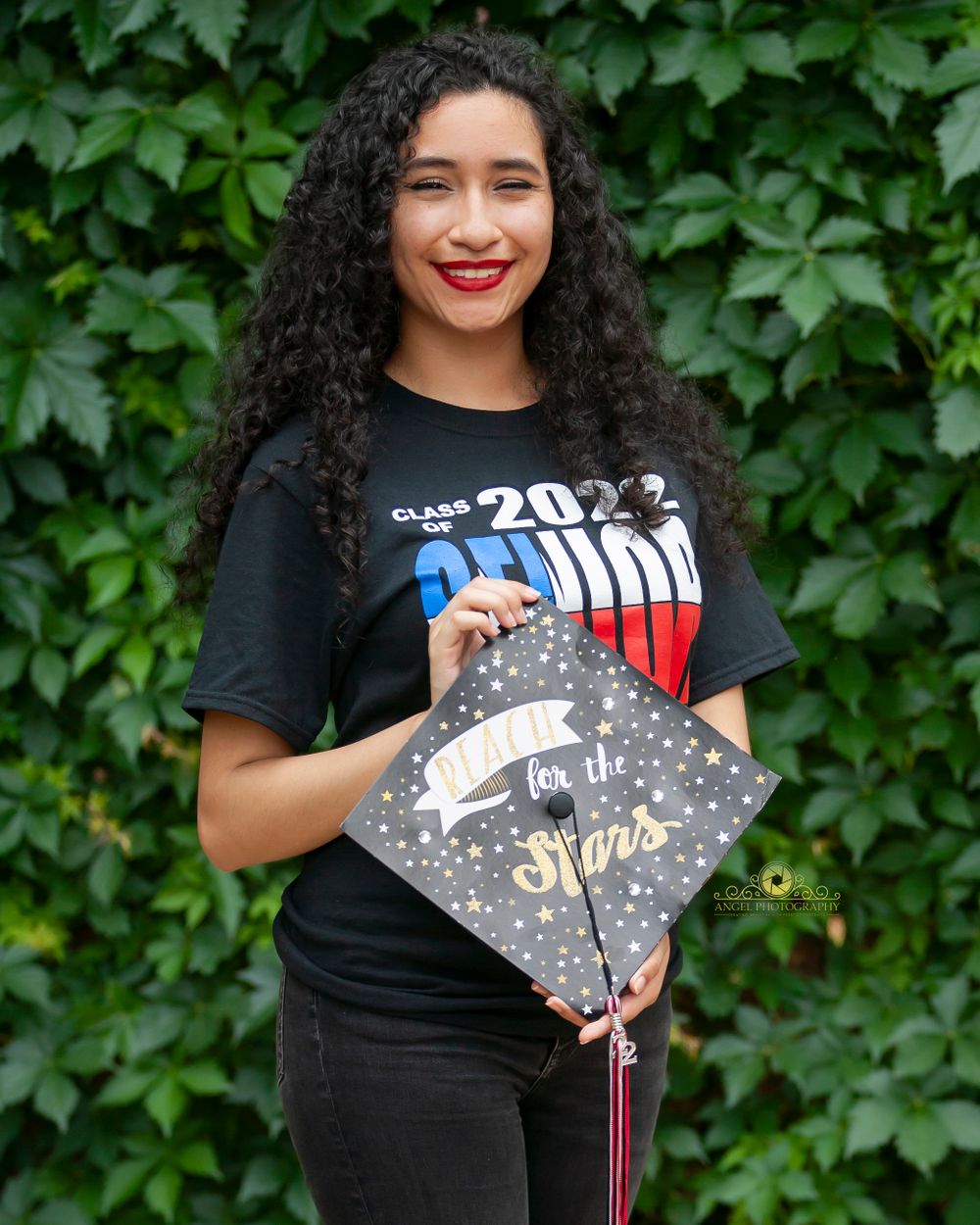 Graduation photo showing decorated mortarboard cap against green leafy background.