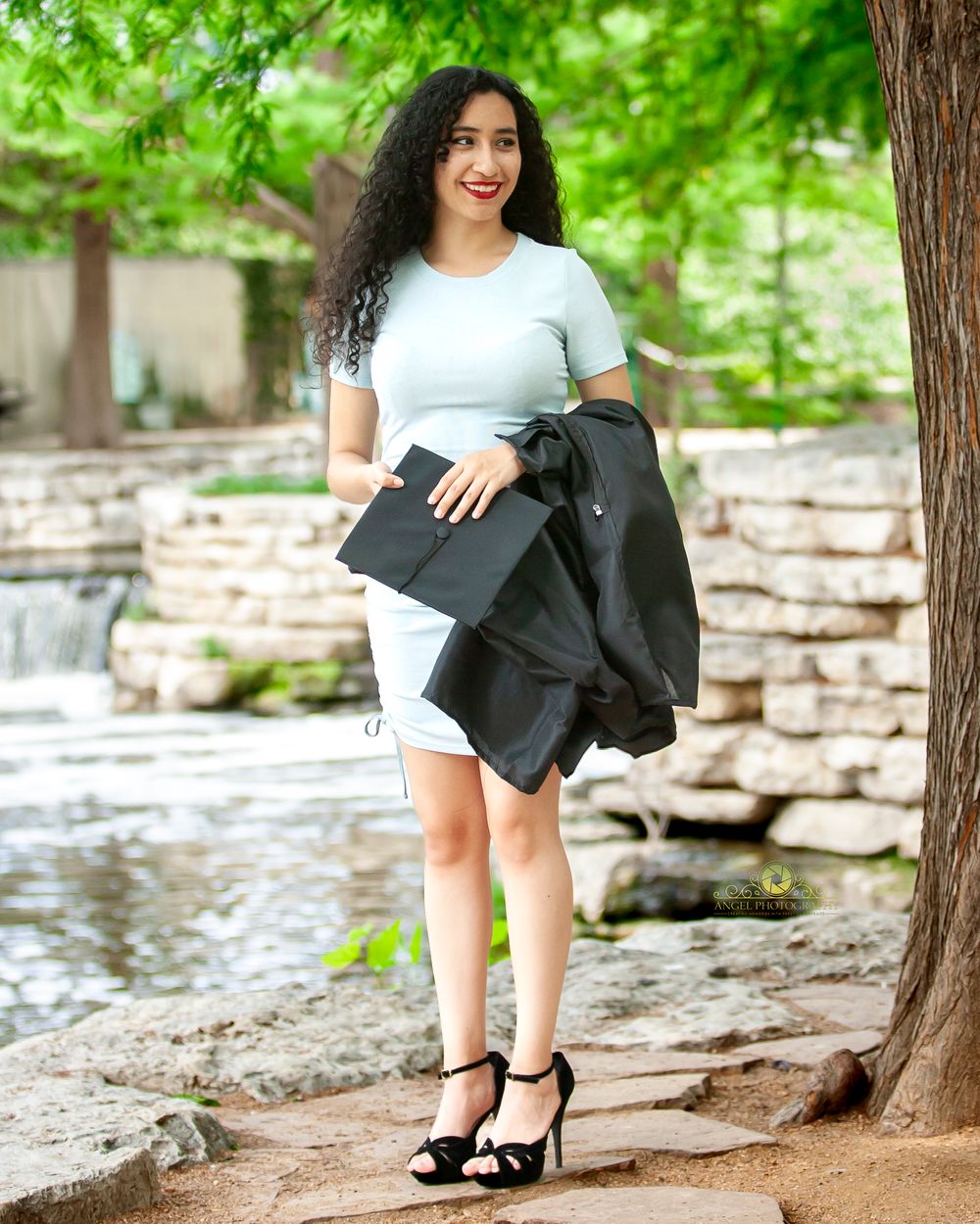 Outdoor portrait in white dress and black jacket near stone waterfall feature.