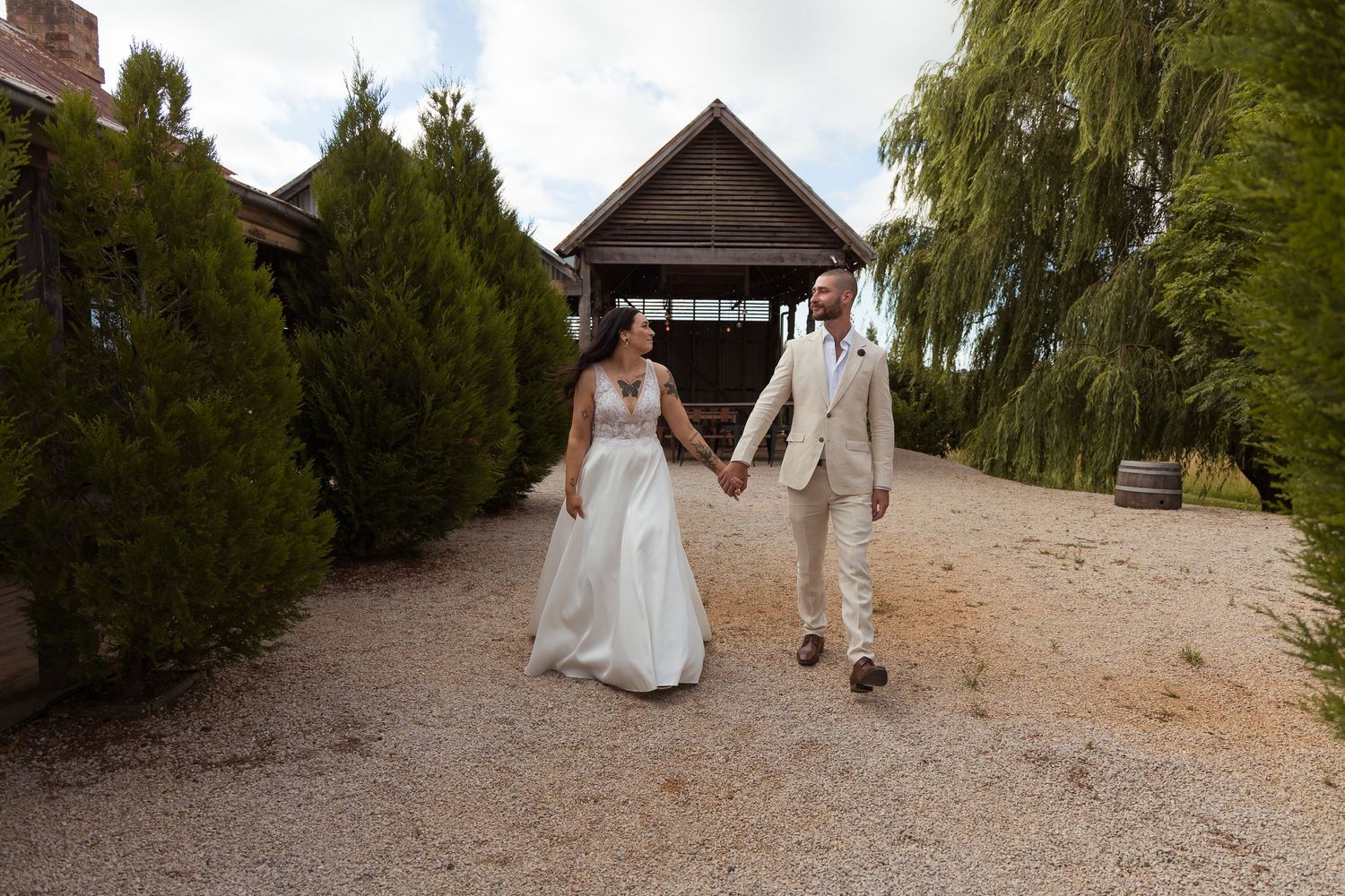Bride and groom holding hands walking along gravel path beside rustic wooden barn venue.