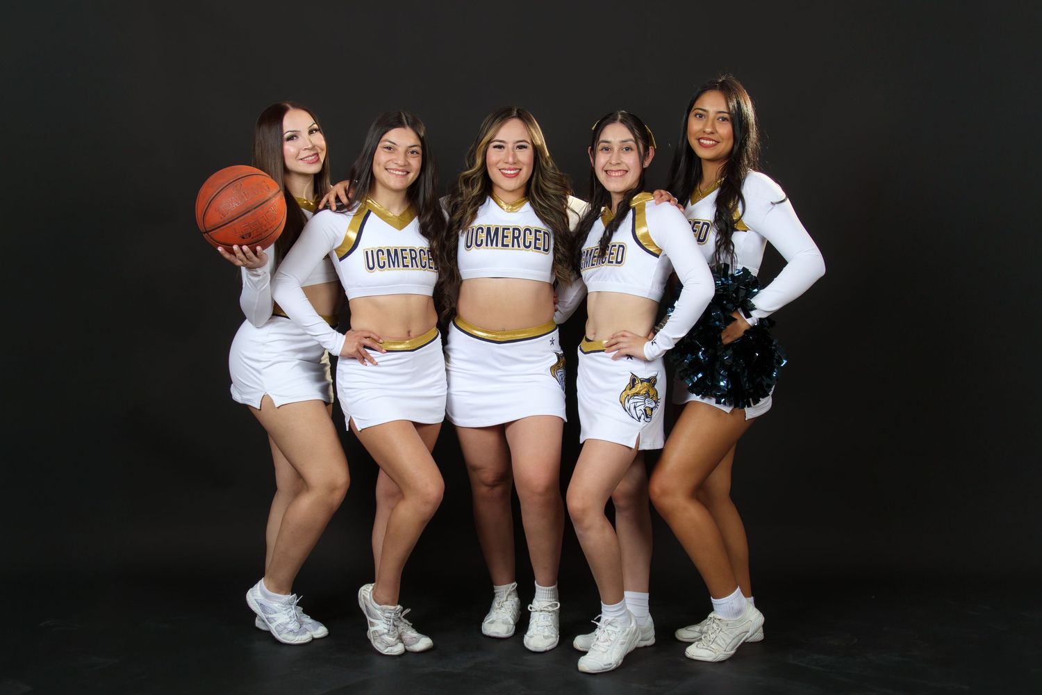 Five UC Merced cheerleaders pose together in white and gold uniforms with a basketball.
