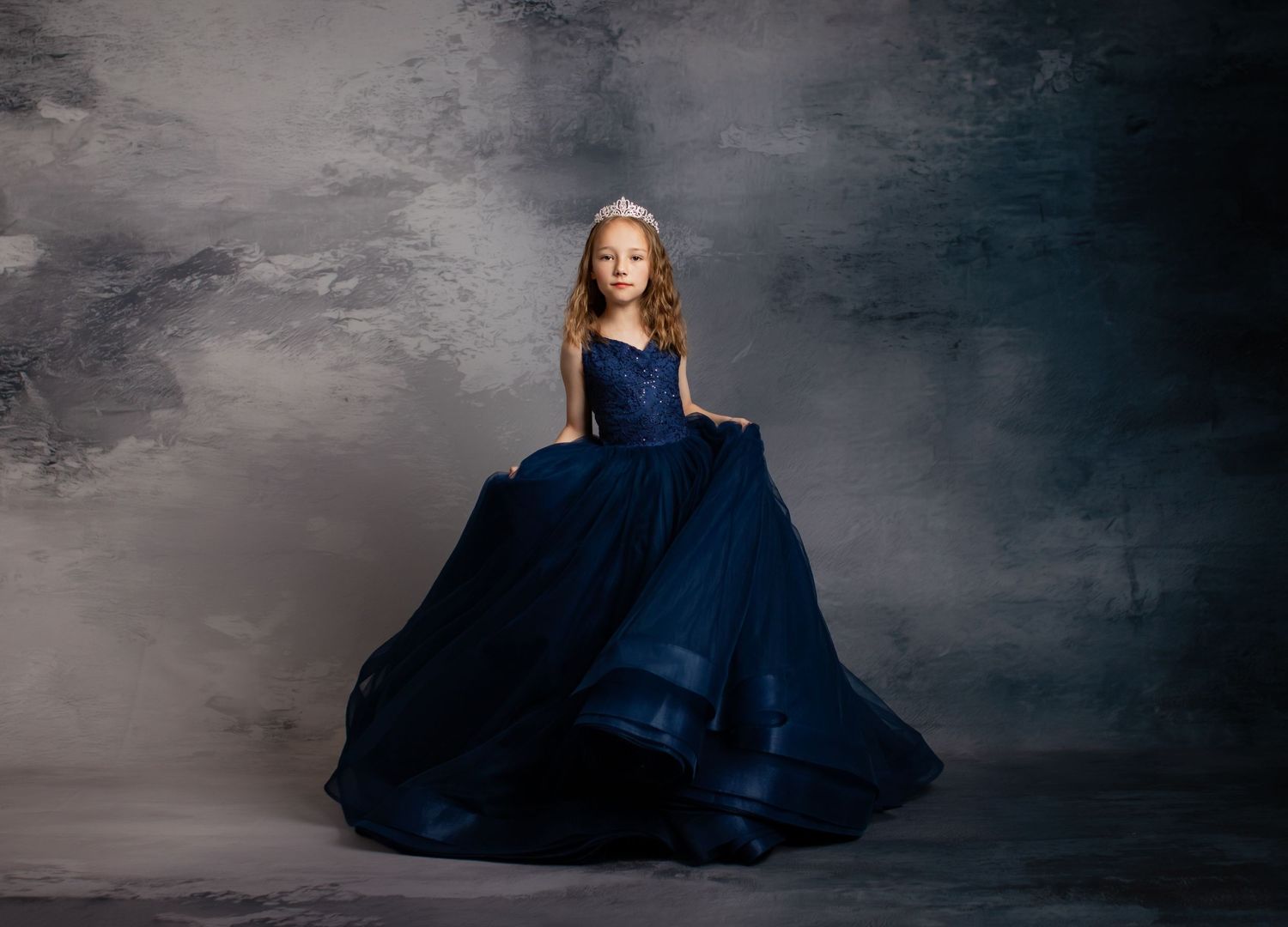 A figure in a flowing navy blue ball gown poses elegantly against a dark textured backdrop.