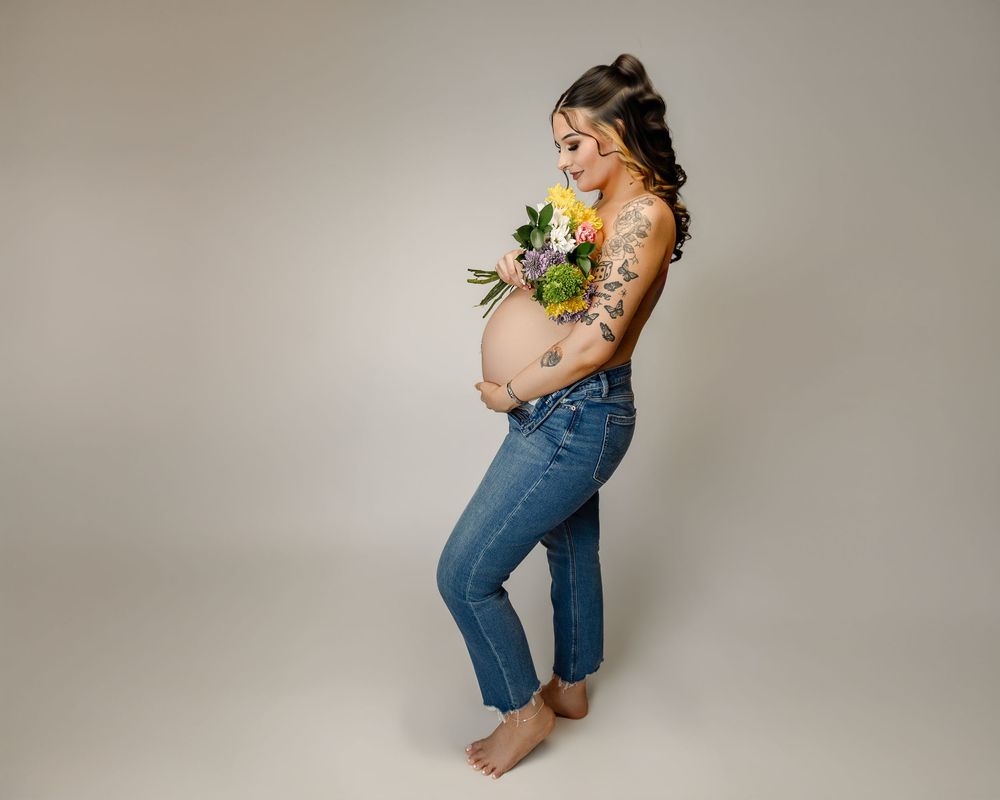 Pregnant woman holding colorful flower bouquet, wearing jeans, showing baby bump, with floral tattoos, studio portrait.