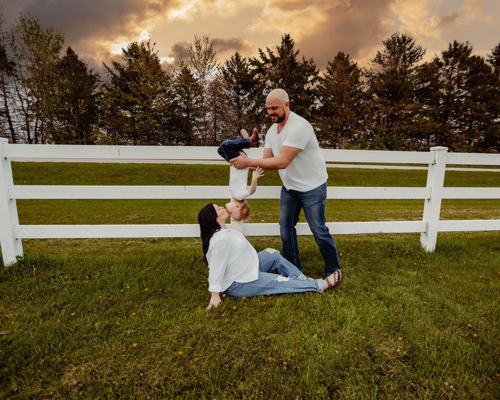 Family playing outdoors on green grass near white fence at sunset, father lifting child while mother sits smiling.
