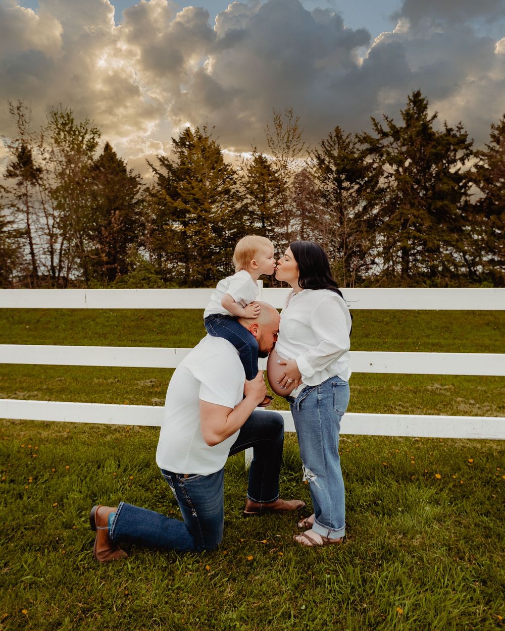 A couple in white shirts kisses their baby outdoors on green grass with a white fence and dramatic sky.