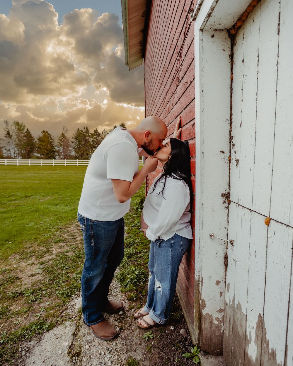 Couple sharing a romantic kiss leaning against a red barn at sunset on a lush green farm.