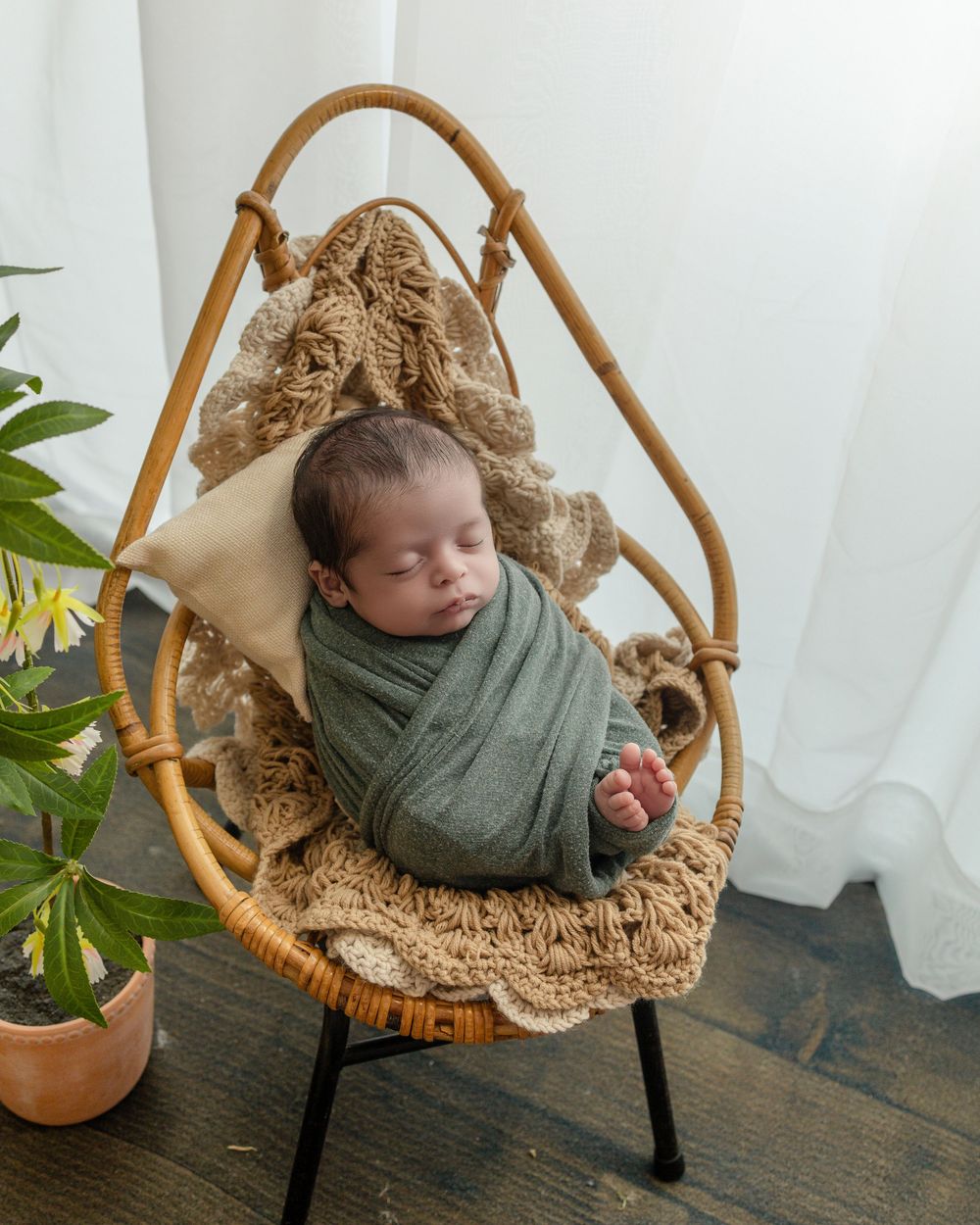 Newborn baby swaddled in green wrap sleeping peacefully in a small rattan chair with crochet blanket.