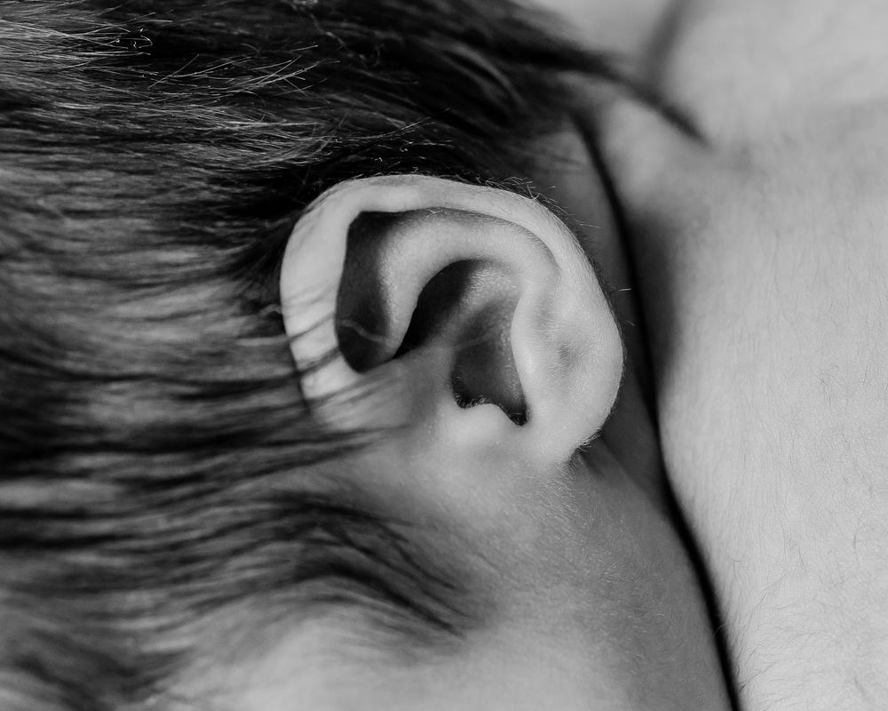 Close-up black and white photo of a human ear with detailed skin texture and dark hair visible in the background.